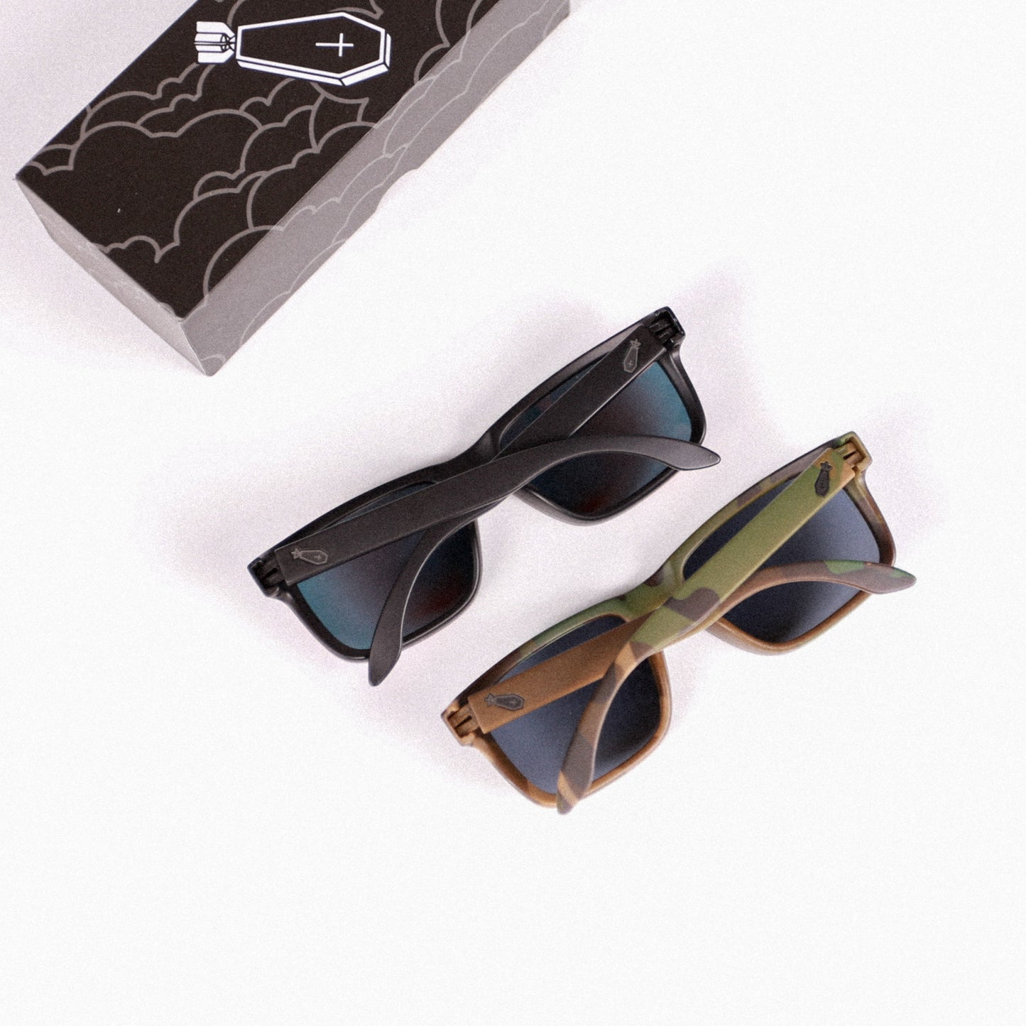 Two pairs of sunglasses, one black and one brown, on a white background with a box featuring a bat design.