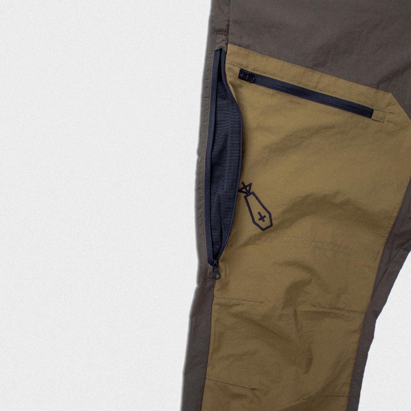 Close-up of a pair of outdoor pants showing a color-blocked design with a dark gray body, a tan panel, and a navy blue zippered pocket.