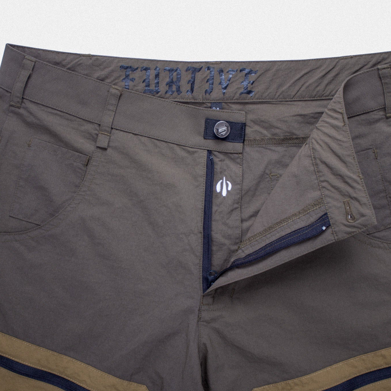Close-up of a pair of technical outdoor pants in a muted gray-green color, featuring tan side panels, dark navy zippers, and a visible 'ZOOM' fabric detail on the waistband.