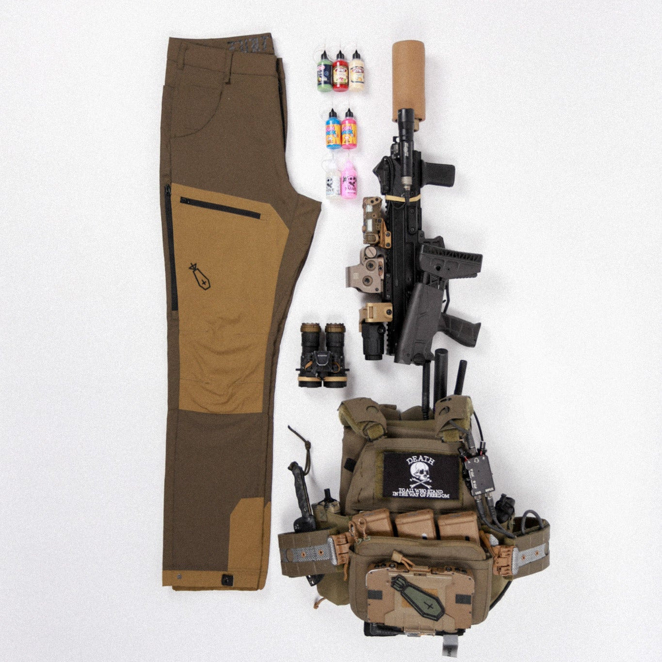 A tactical gear flat lay on a white background, featuring a pair of brown and tan pants, a black rifle with a tan stock, a tan utility vest, and small accessories including a pair of sunglasses and a small bottle.